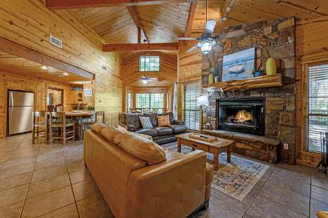 Cozy wooden cabin interior with a stone fireplace, leather sofas, and a dining area in the background.