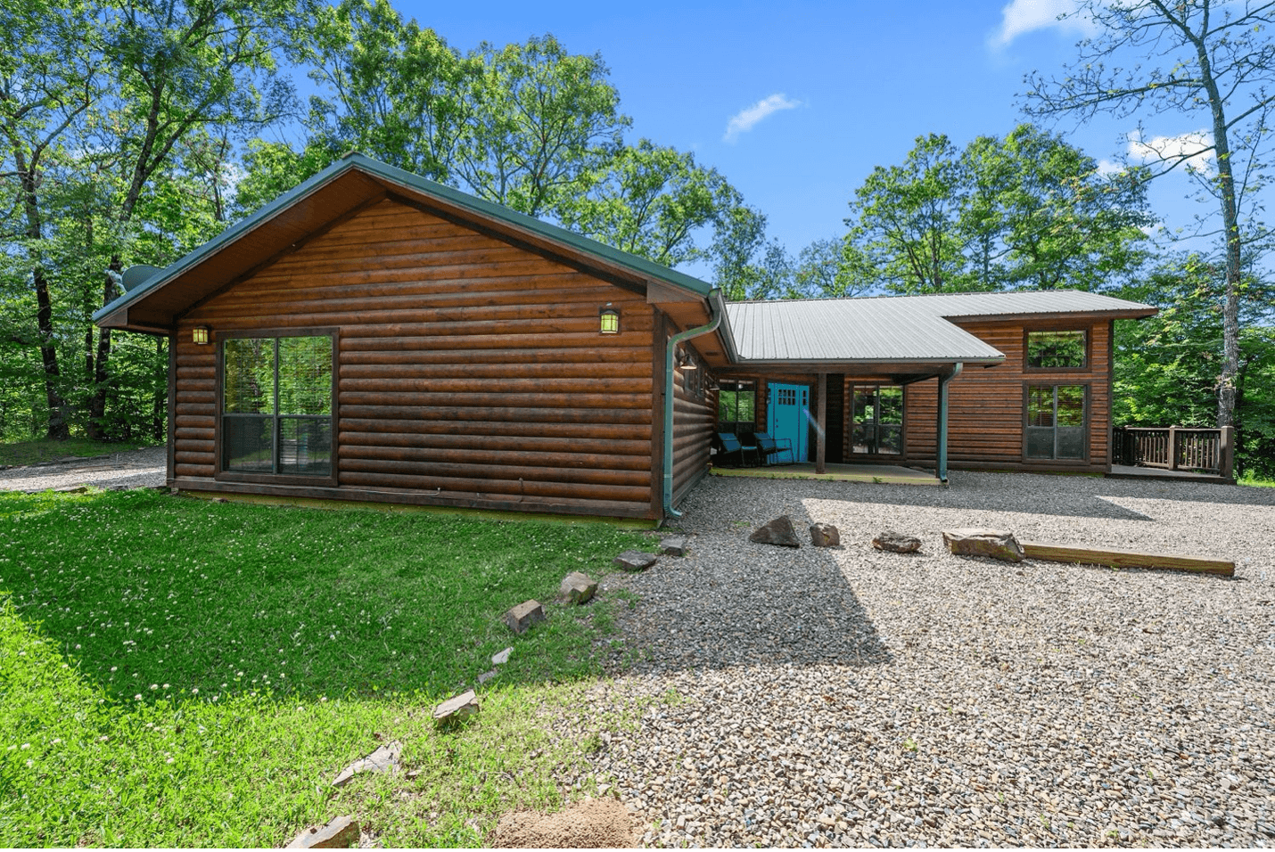 A log cabin with a green roof amidst trees, featuring a gravel driveway and green lawn.