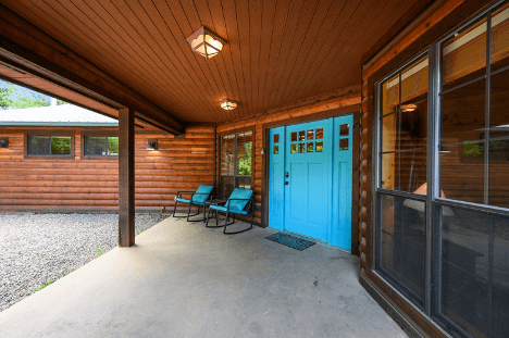 Front porch of a log cabin with a bright blue door and two black chairs.