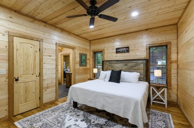 Cozy rustic bedroom with wood walls, a white bed, ceiling fan, and a patterned area rug.