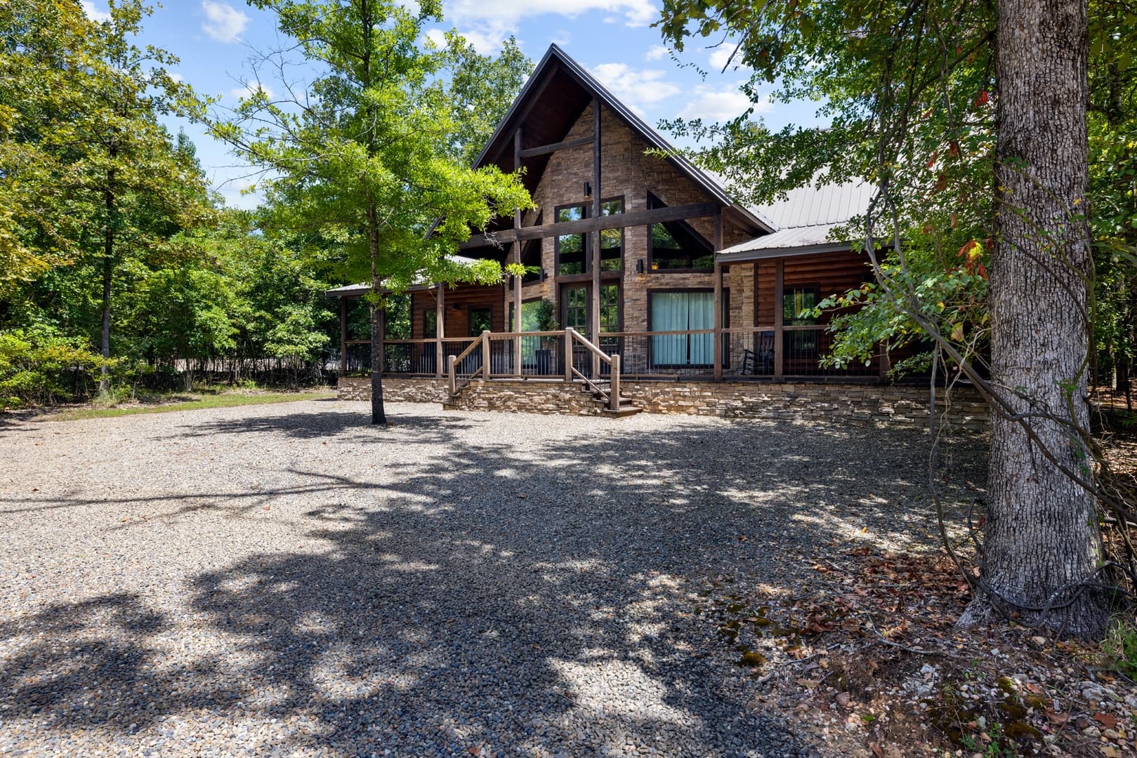 A rustic log cabin with a large gable, front porch, and gravel driveway amidst trees.
