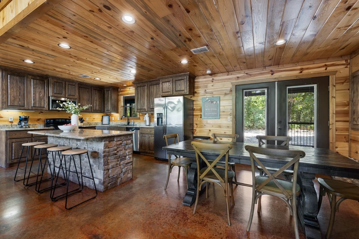 Rustic wooden kitchen interior with stone breakfast bar and dining table, leading to a glass door.