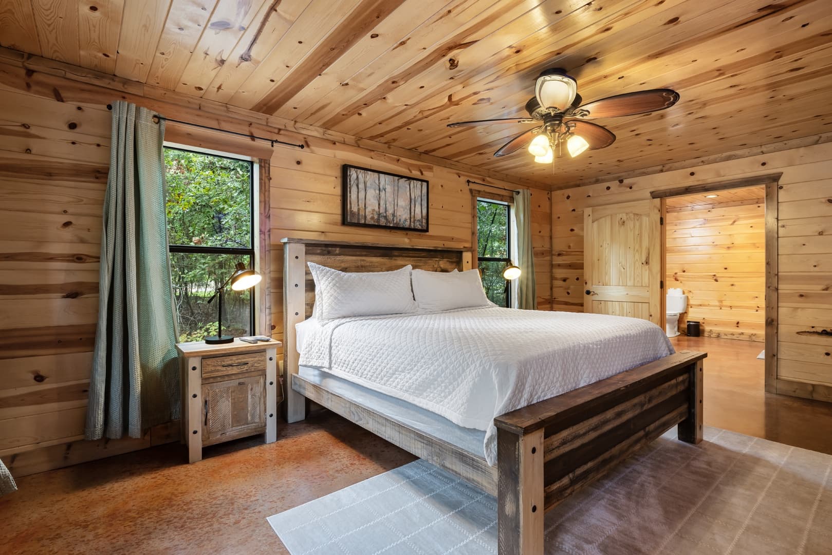 Cozy wooden cabin bedroom with a rustic bed, side tables, ceiling fan, and forest view window.