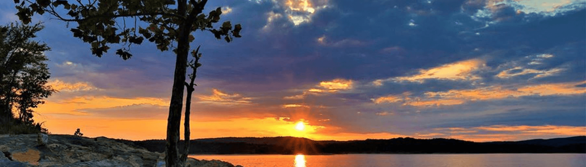 Sunset over a calm lake, framed by silhouetted trees and colorful clouds.