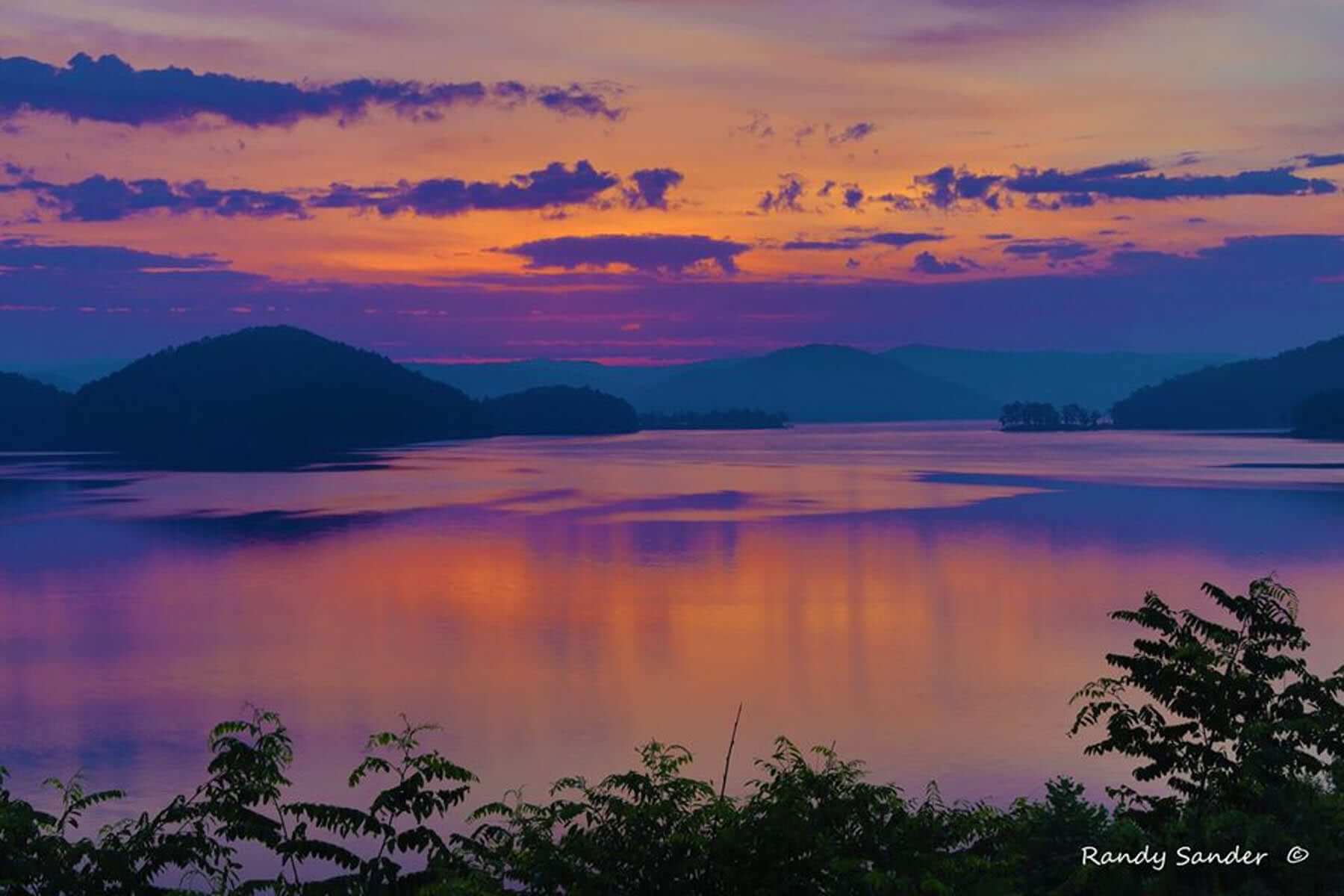 A tranquil lake reflects a vibrant sunset with hills and trees framing the scene.
