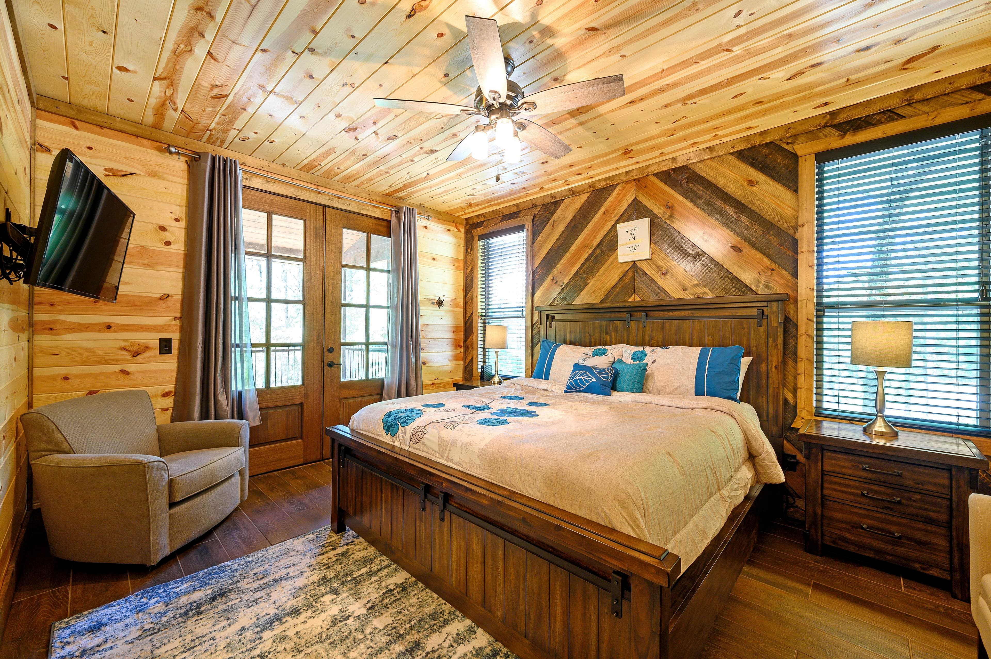 A cozy bedroom with wooden walls, a large bed, a seating area, and natural light from windowed doors.