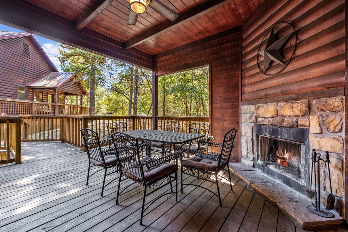 A wooden deck featuring a dining table and chairs beside a stone fireplace, surrounded by trees.