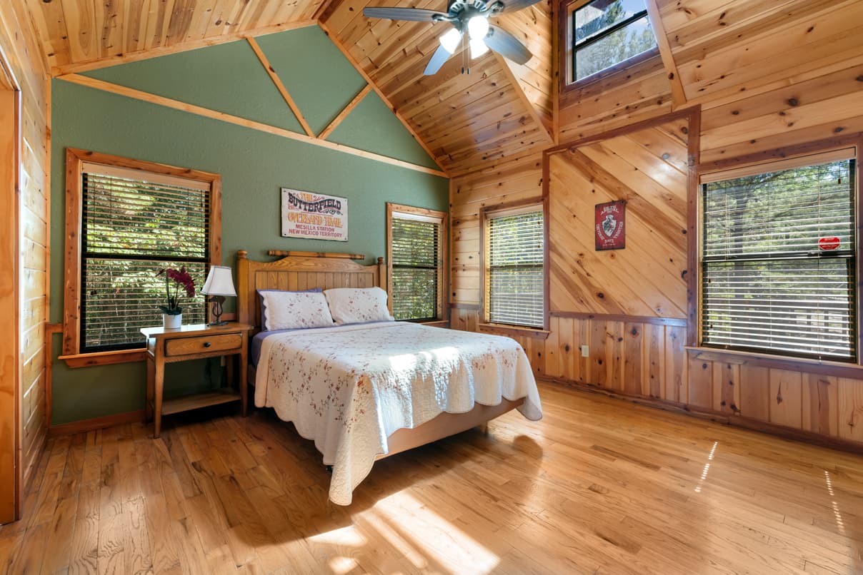 A cozy wood-paneled bedroom featuring a queen bed, bedside table, and large windows letting in natural light.