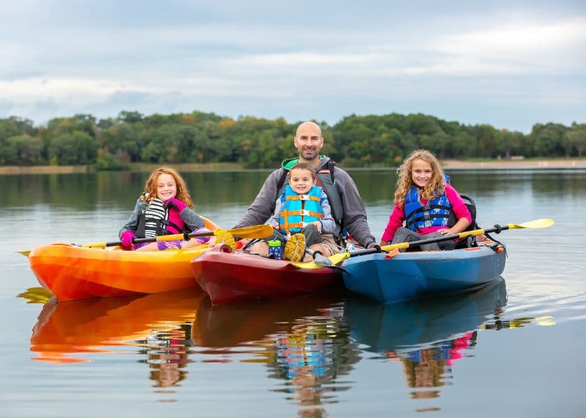 A man and three children sit in colorful kayaks on a calm lake, surrounded by trees under a cloudy sky. A man and three children sit in colorful kayaks on a calm lake, surrounded by trees under a cloudy sky.