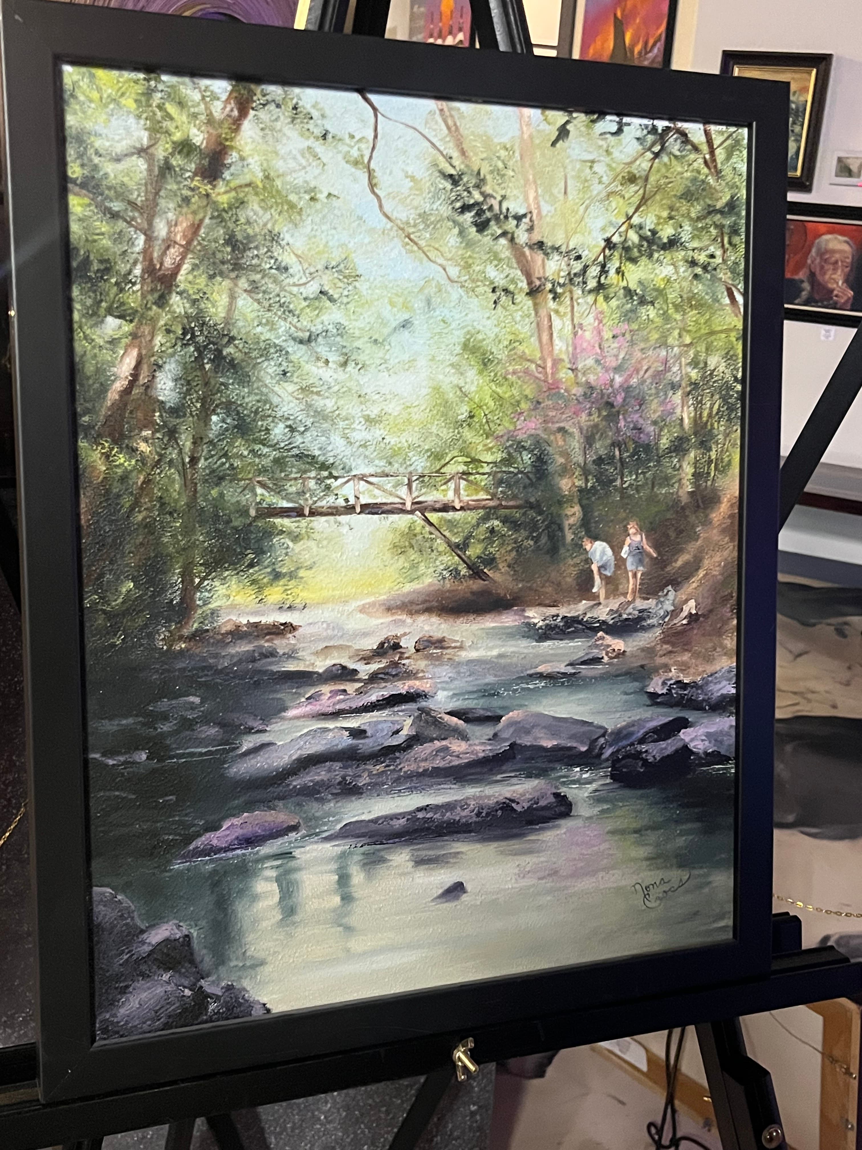 A framed painting depicting a serene landscape with a stream, rocks, and two people near a small wooden bridge.