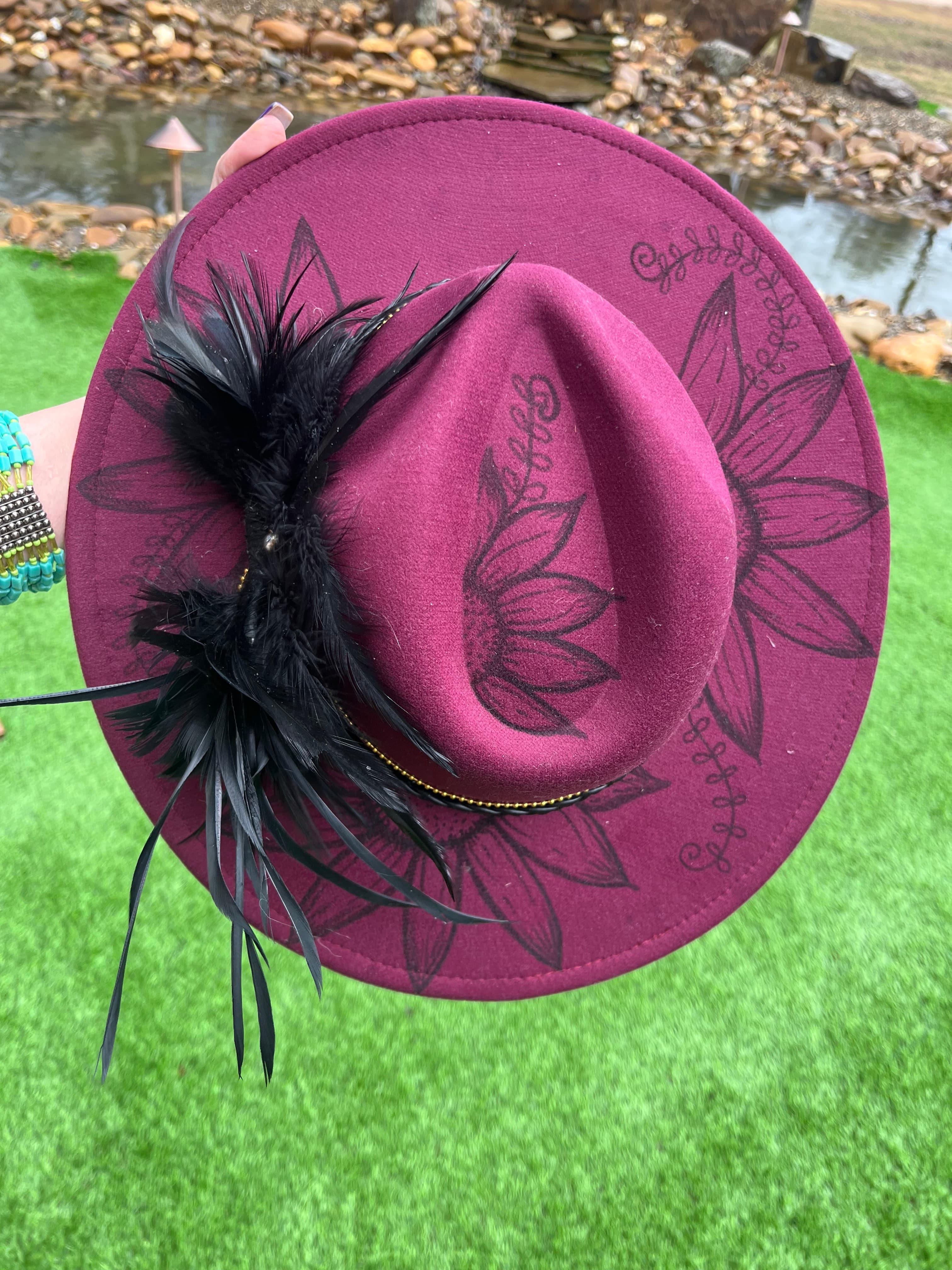 A maroon hat adorned with black feathers and floral engravings is held against a grassy background.