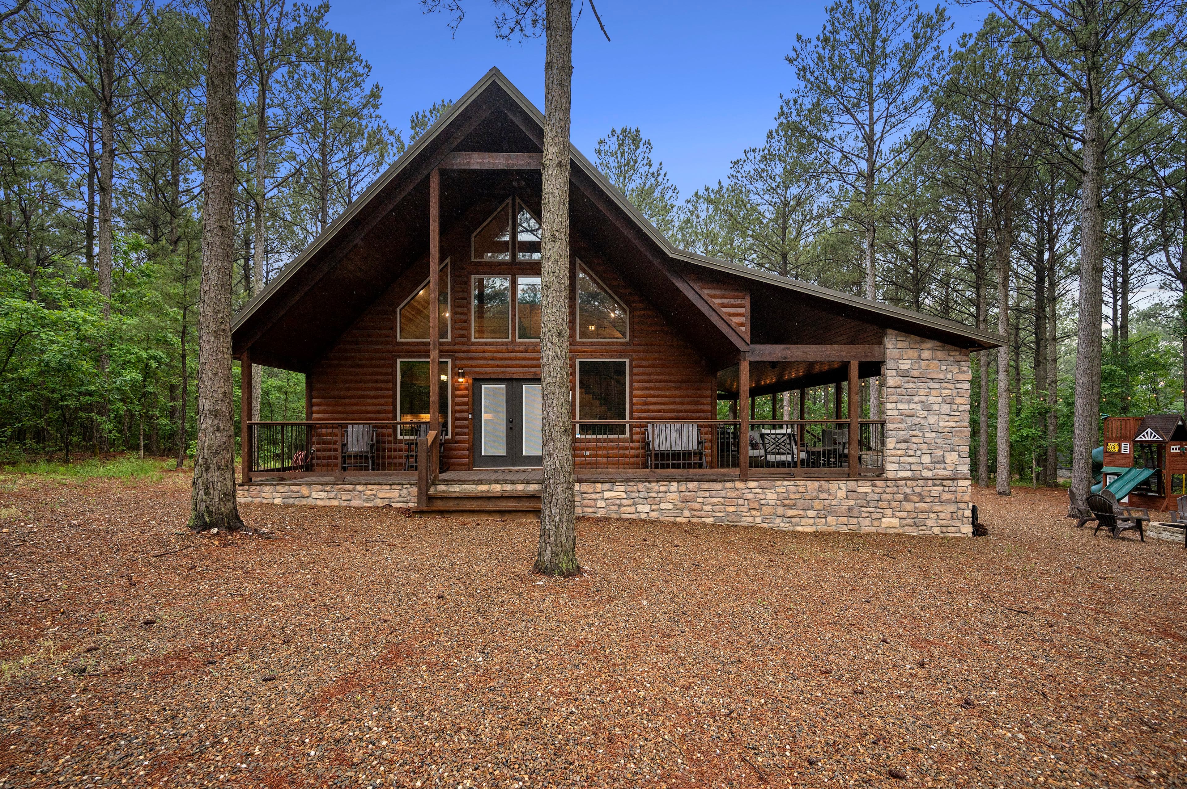 A rustic wooden cabin surrounded by tall trees and gravel ground.