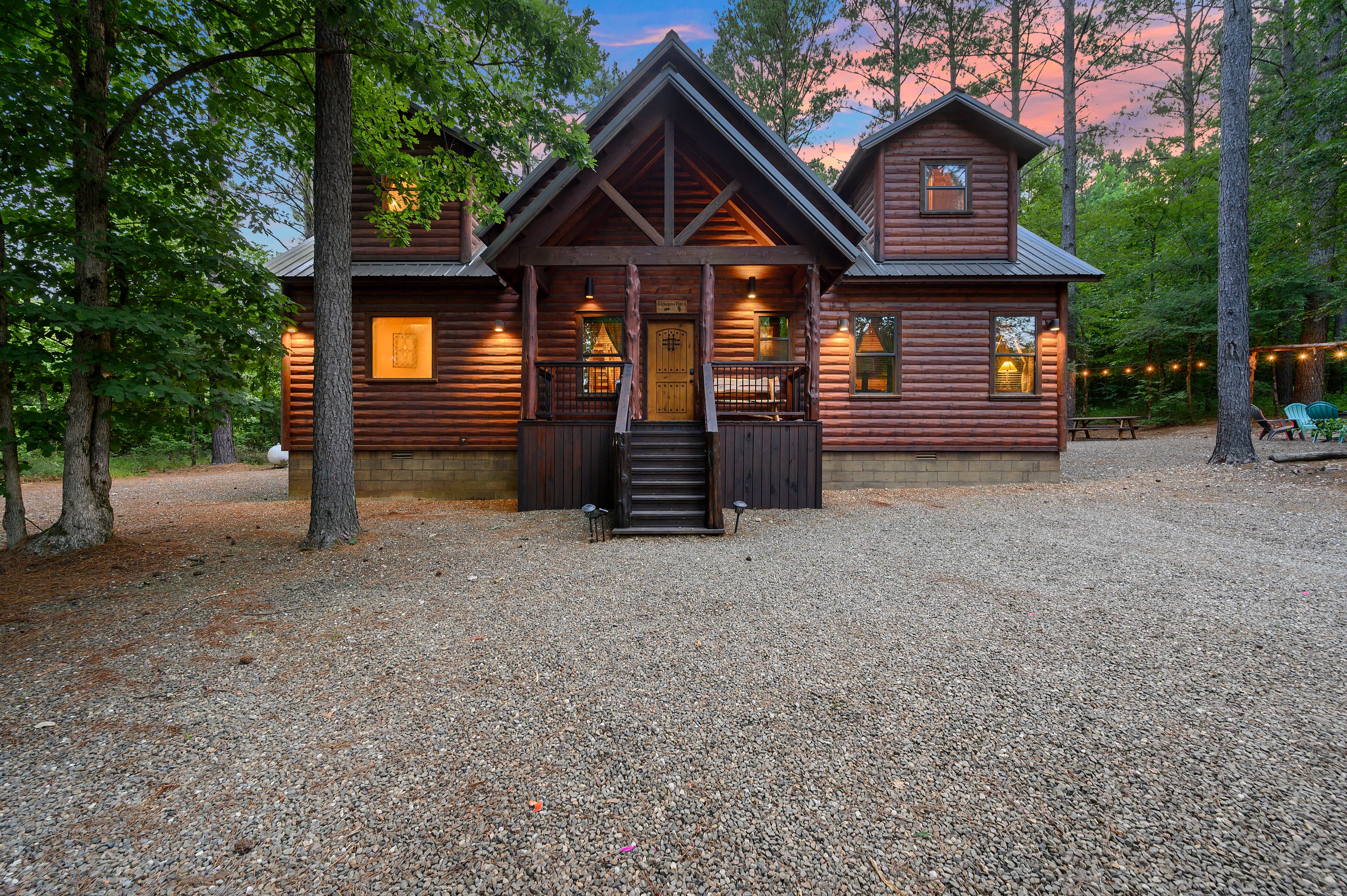 A charming log cabin surrounded by trees, with a gravel driveway and warm evening lighting.