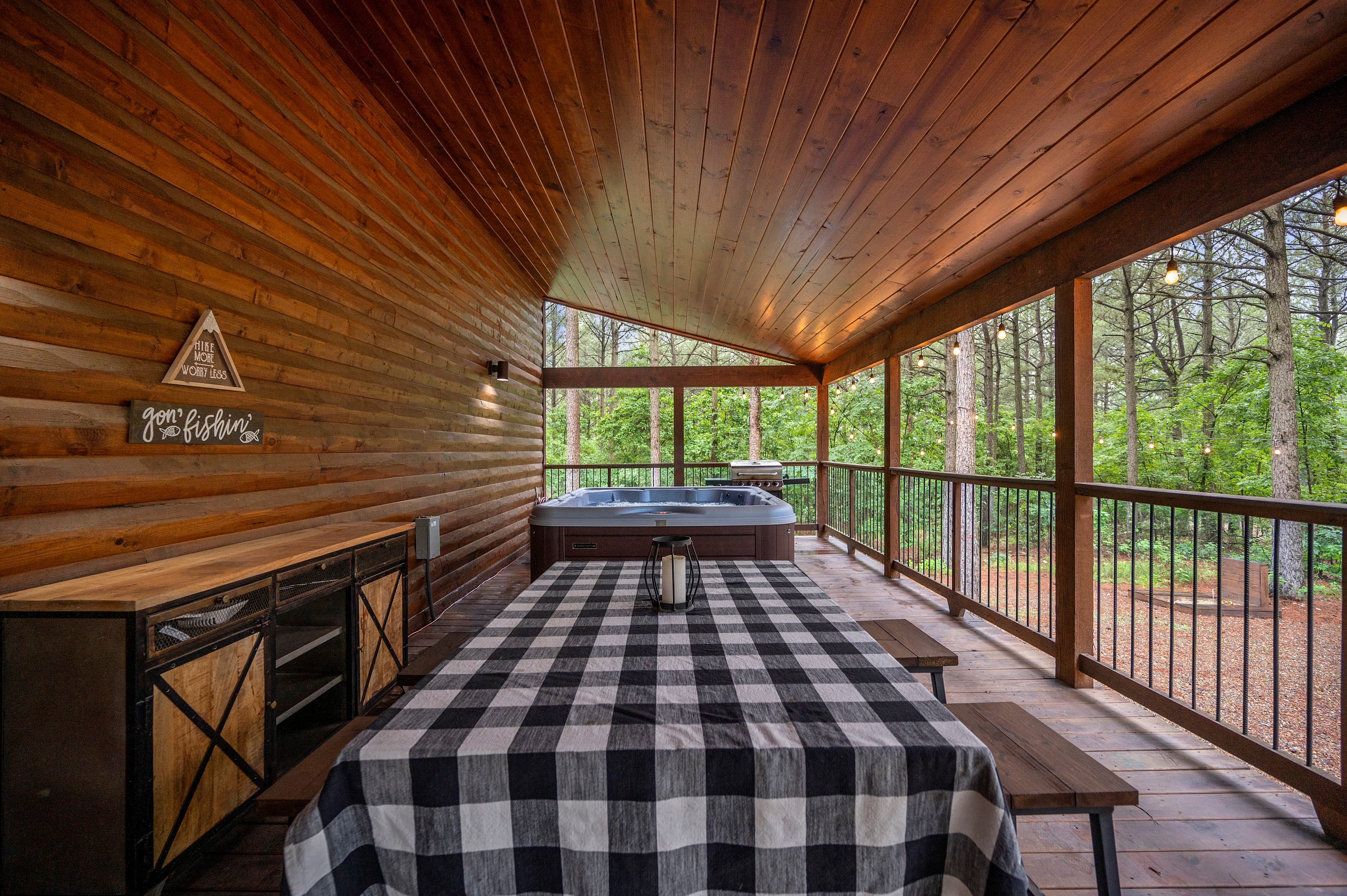 A cozy wooden deck featuring a picnic table with a checkered tablecloth and a hot tub, surrounded by lush greenery.