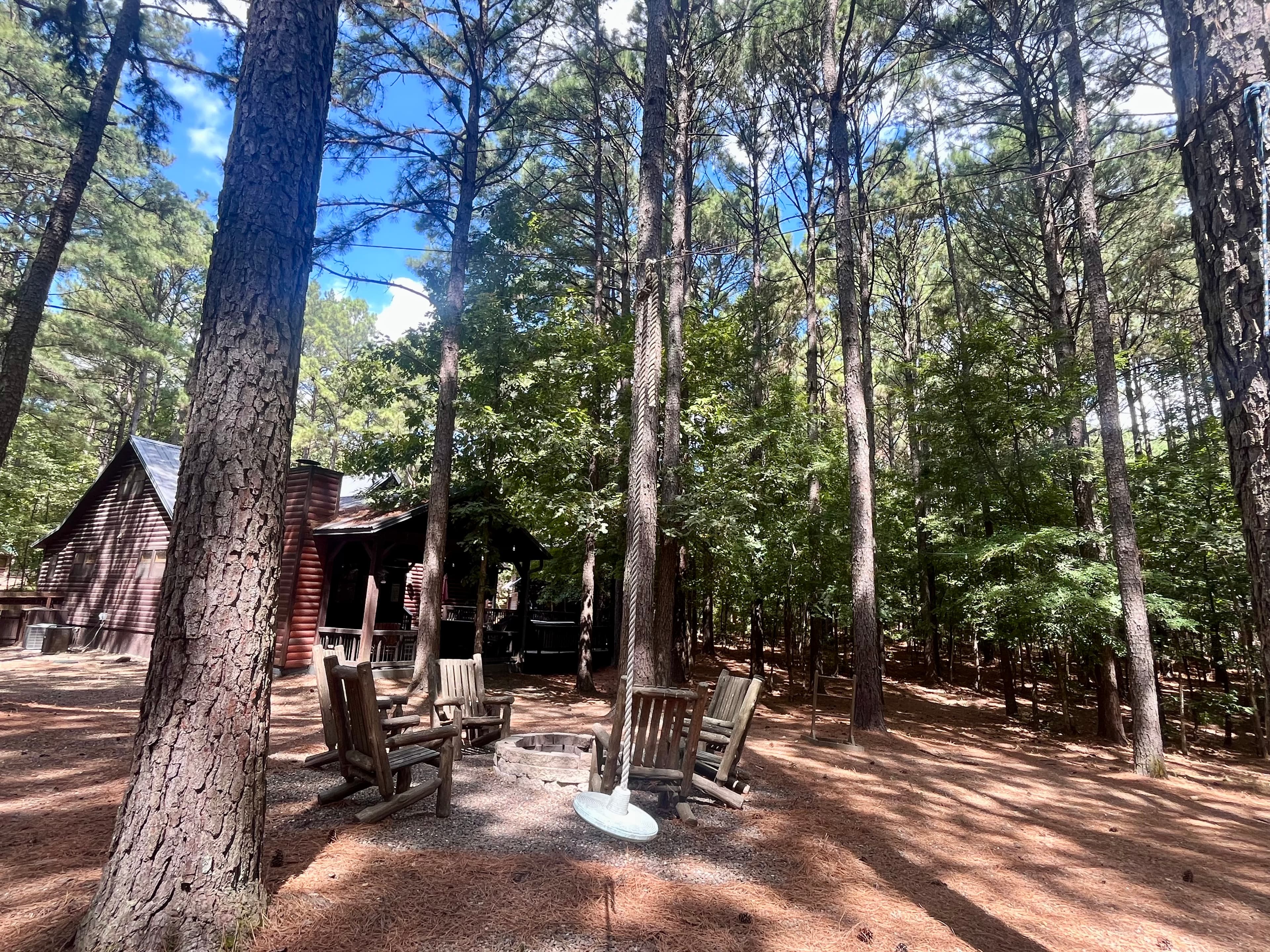 A rustic cabin surrounded by tall pine trees, featuring a fire pit and wooden seating area.