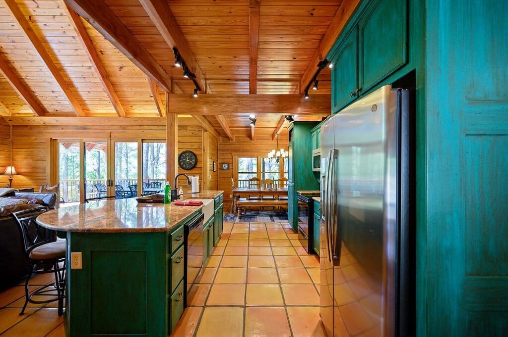 A spacious rustic kitchen with green cabinetry, wooden beams, and tile flooring, featuring a stainless steel refrigerator and a view of an outdoor dining area.