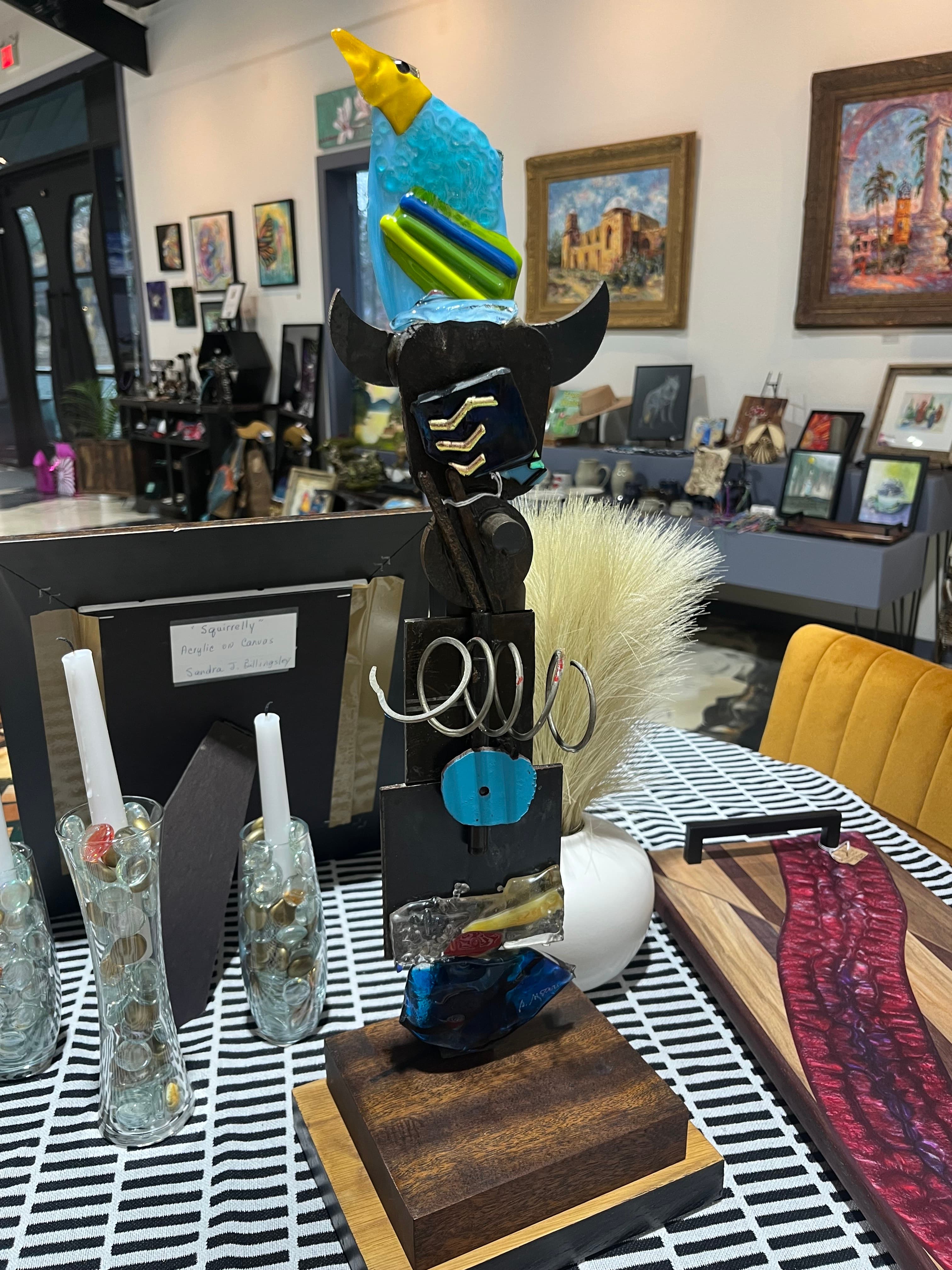 A colorful bird sculpture perched on a metallic base, surrounded by decorative glass vases and artwork in a gallery setting.