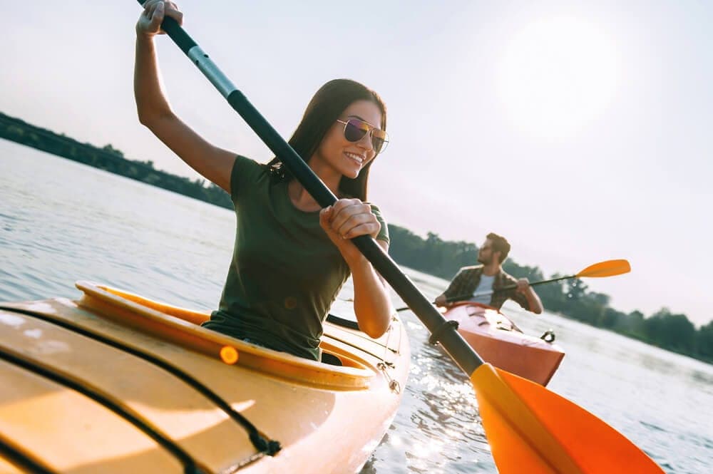 Two people are kayaking on a sunny day, with one woman smiling as she paddles.