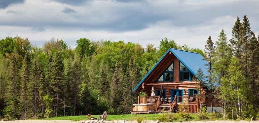 A log cabin with a blue roof surrounded by lush green trees under a cloudy sky.