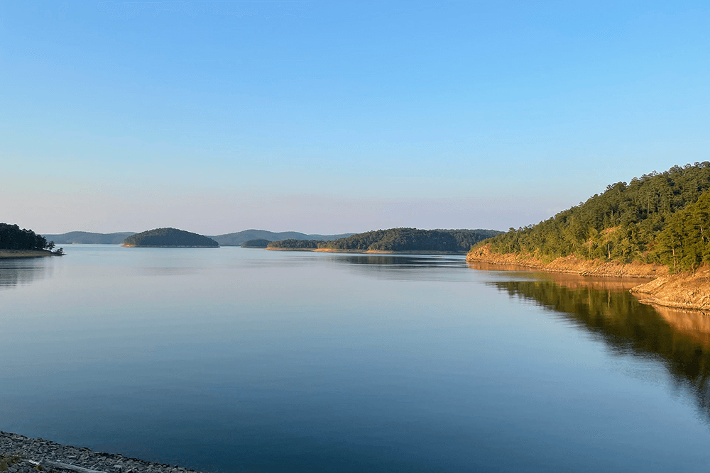 A calm lake reflecting blue skies and surrounded by forested hills.