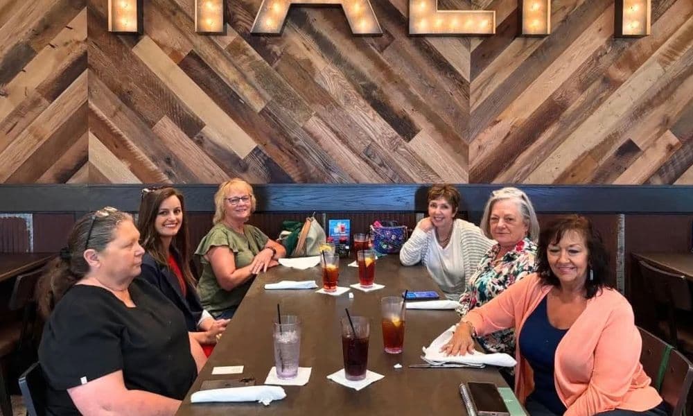 Six women smile around a long wooden table at a restaurant in Broken Bow, Oklahoma, with iced drinks in front of them.