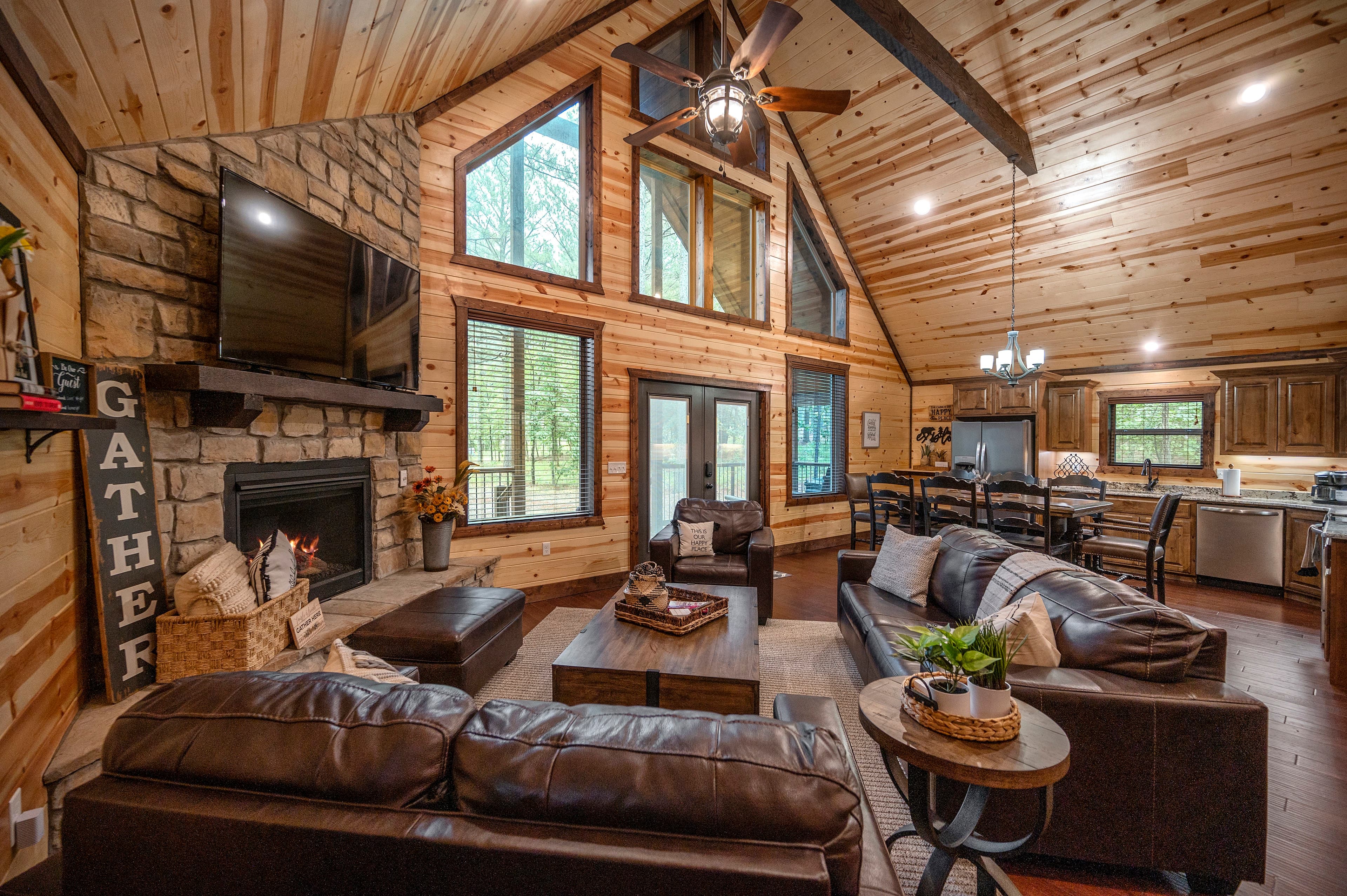 A cozy living room with leather sofas, a stone fireplace, large windows, and wooden walls.