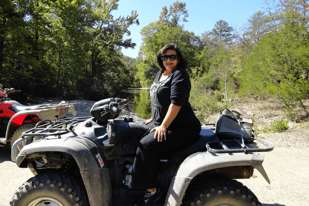 A woman in sunglasses sits on an ATV beside a calm stream surrounded by trees.