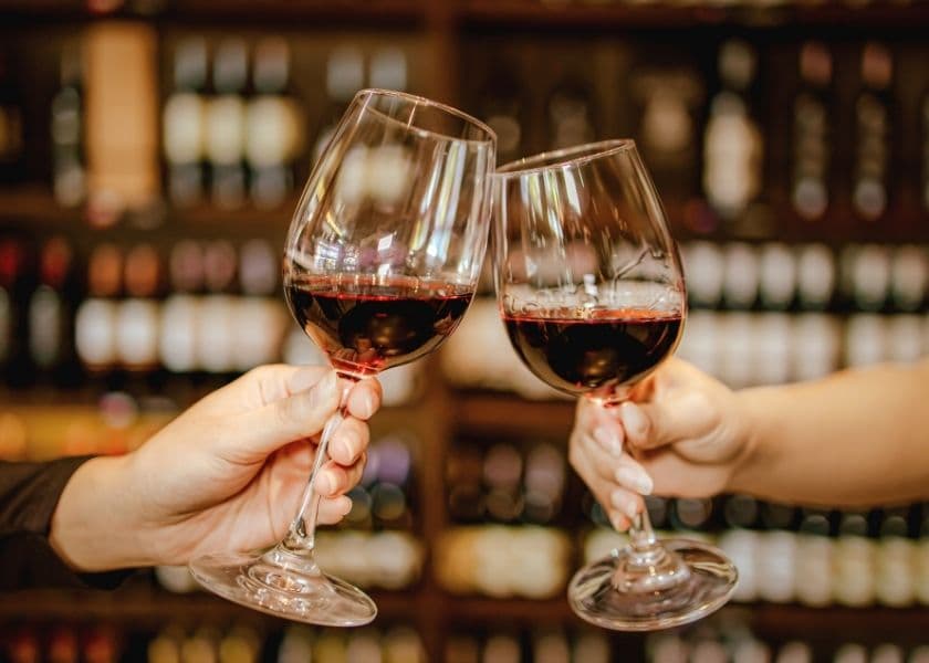 Two hands clink glasses of red wine in a cozy setting with a wine shelf in the background.