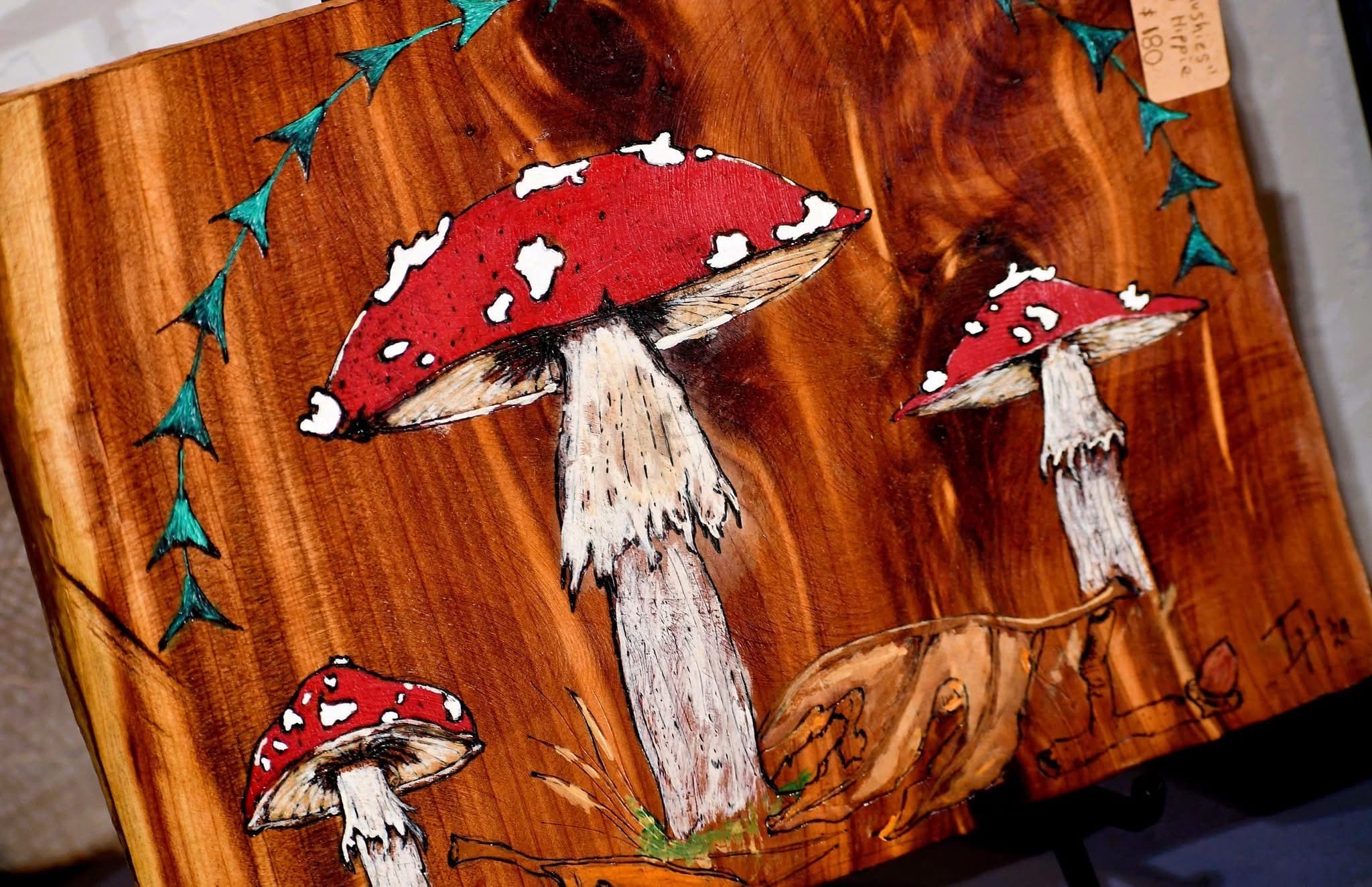 A wooden artwork featuring three red mushrooms and green vines intricately painted.