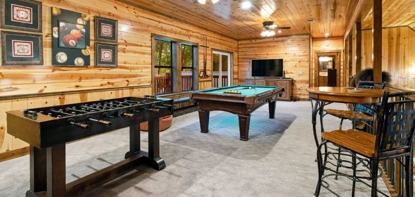 A wood-paneled game room featuring a foosball table, a pool table, and a small bar area.
