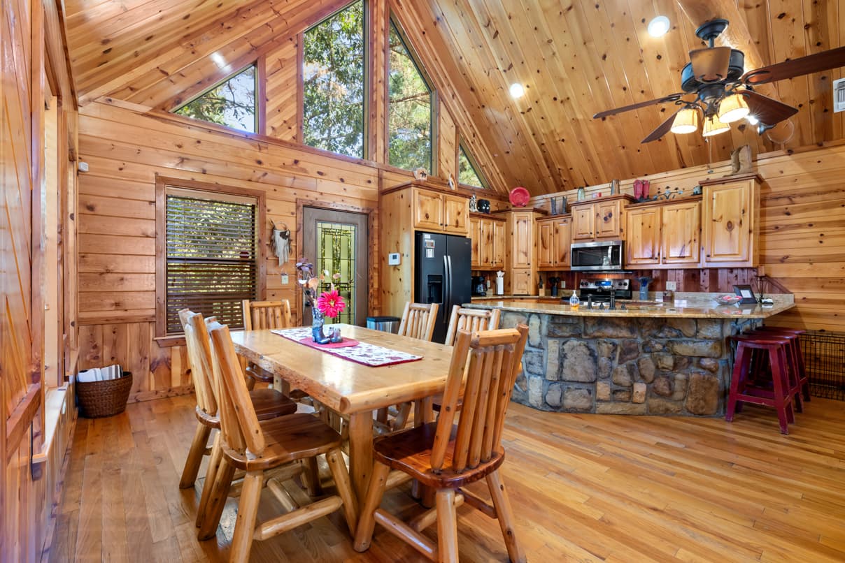 A cozy wooden dining area with a table and chairs, leading into a kitchen with stone accents and large windows.