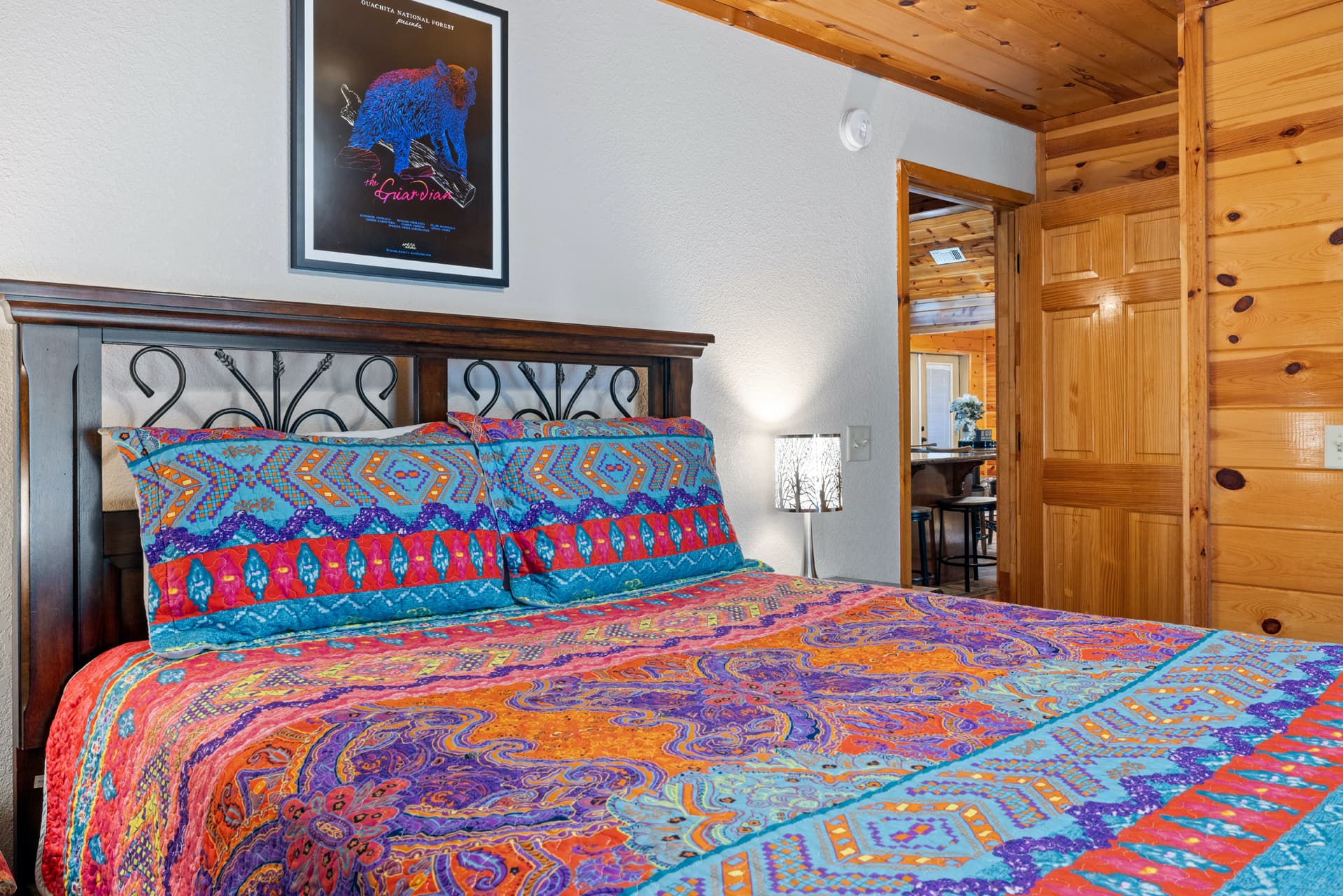 A vibrant, patterned quilt covers a bed in a cozy wooden room, with a decorative lamp and a colorful bear-themed poster on the wall.