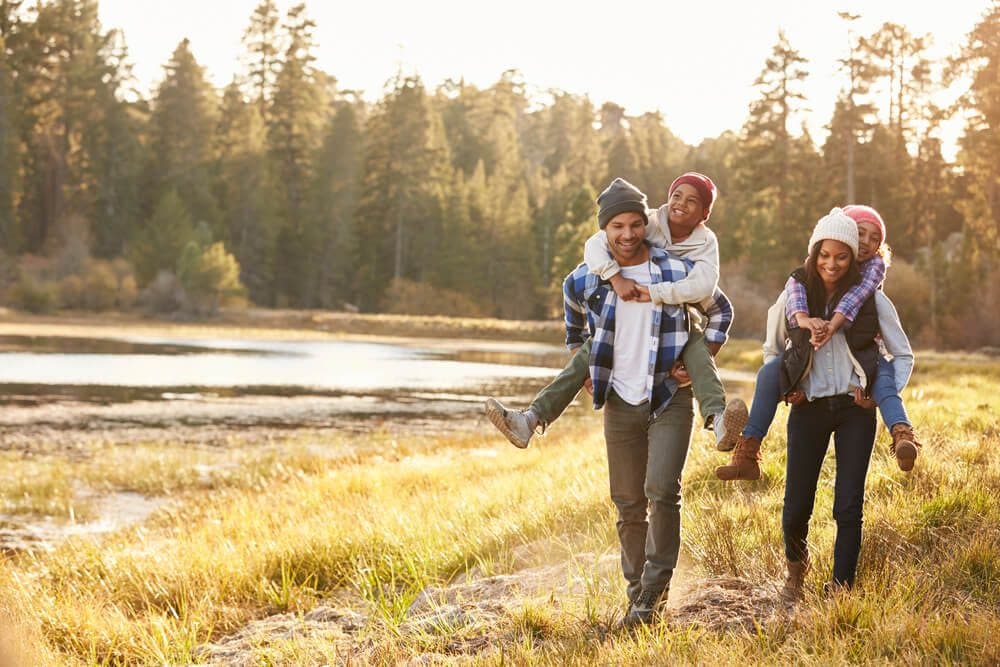 A group of four people enjoys a playful outing in nature, with two children riding on the backs of adults near a lake surrounded by trees.