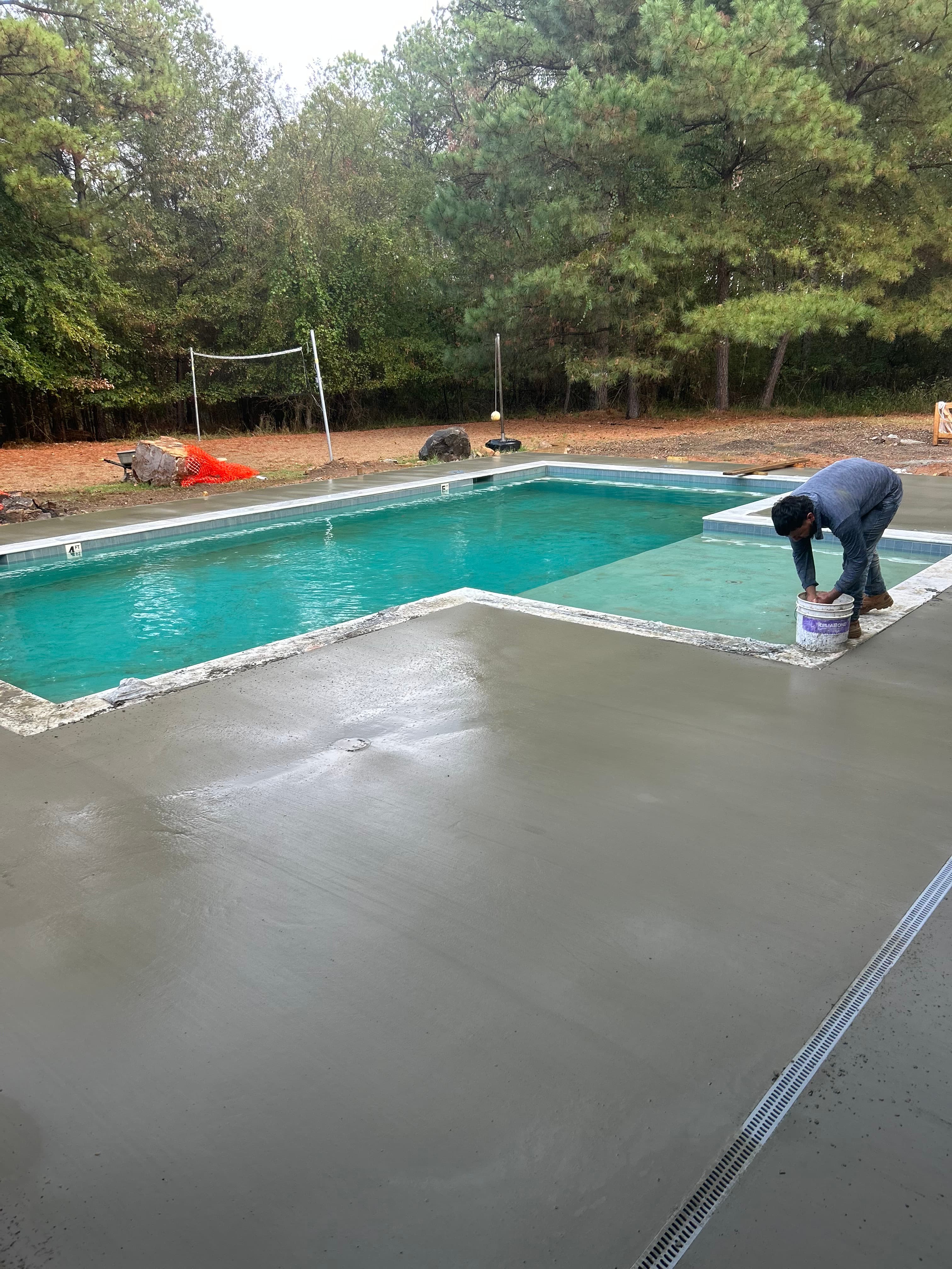 A person is painting the edge of a swimming pool with a bucket of paint, surrounded by trees.