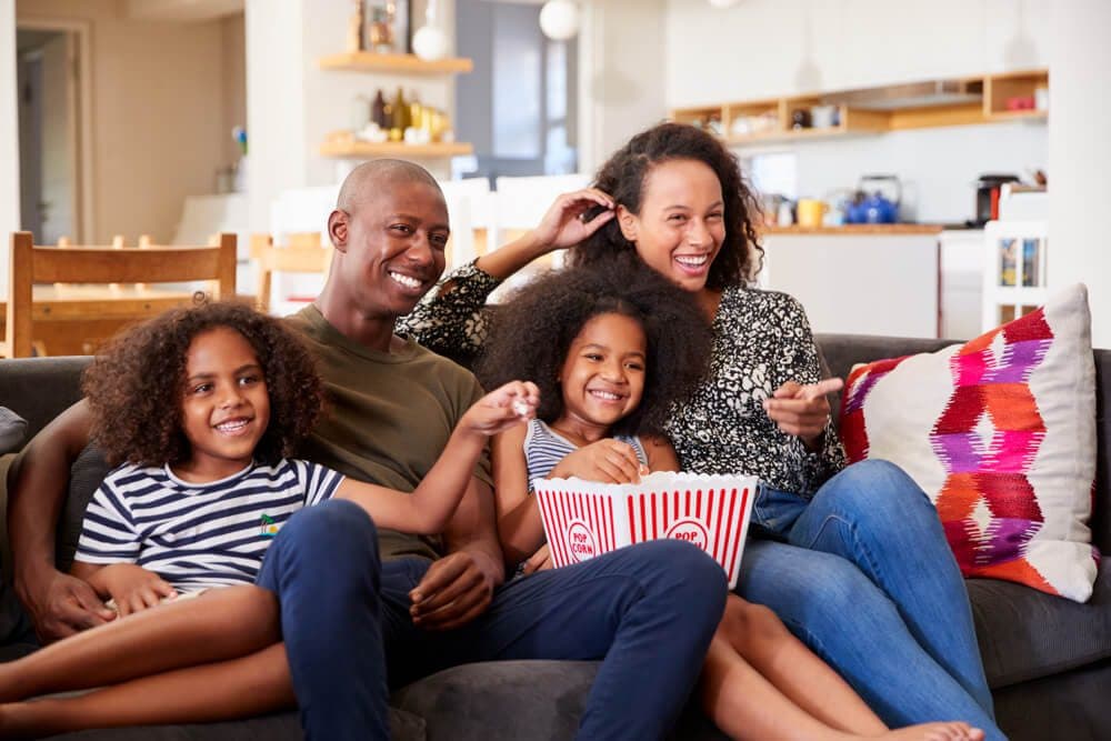 A smiling family of four enjoys a movie night on the couch with popcorn.