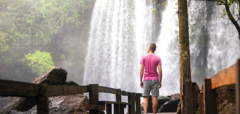 A person stands on a wooden path, gazing at a cascading waterfall surrounded by lush greenery.