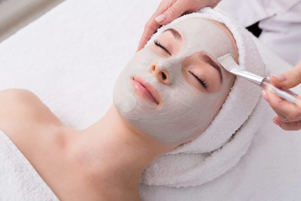 A woman receives a facial treatment with a green clay mask applied to her face.
