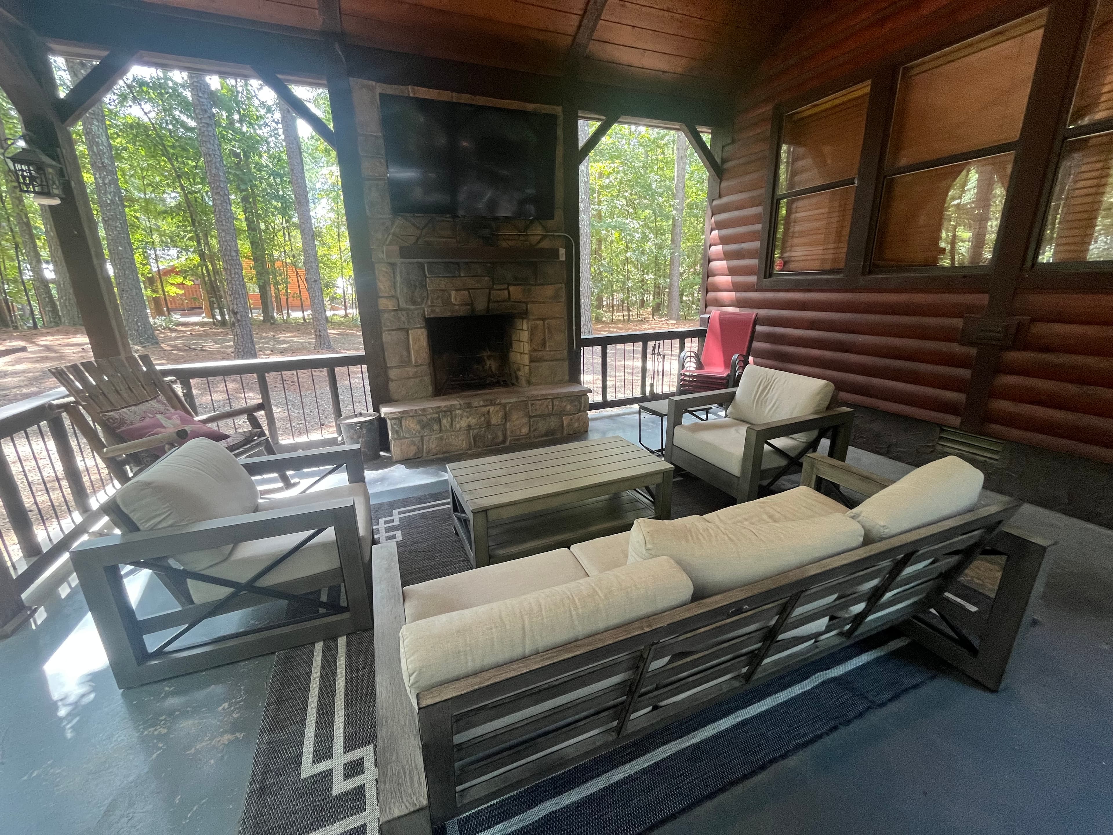 Cozy outdoor seating area with a stone fireplace and TV, surrounded by trees.