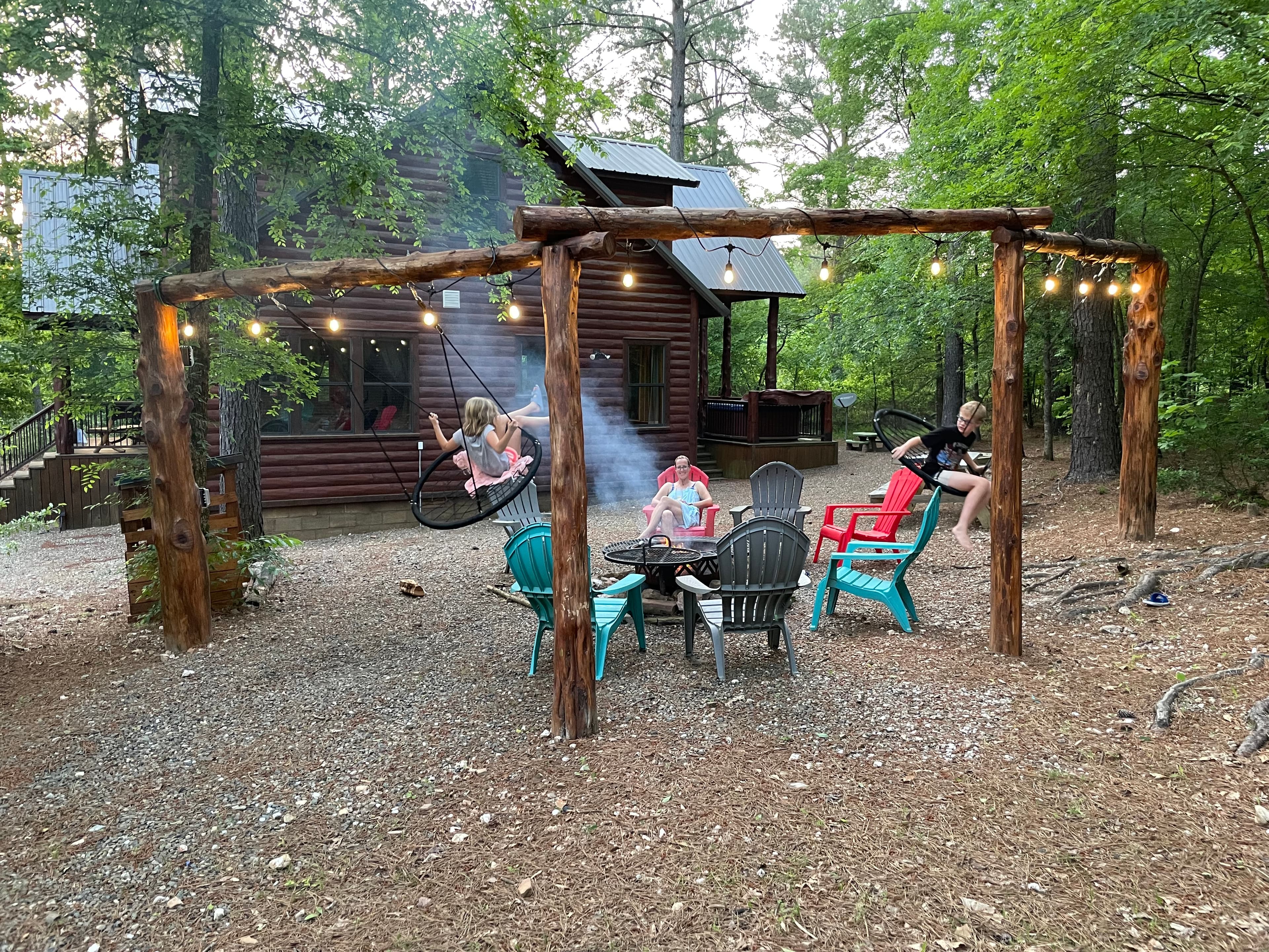 A cozy outdoor seating area with colorful chairs and a fire pit in front of a log cabin surrounded by trees.