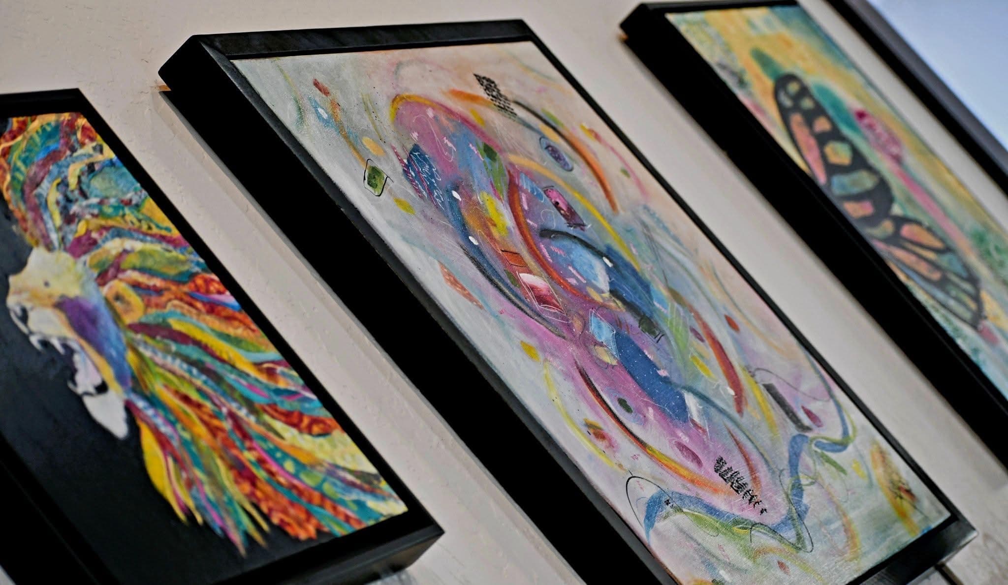 Three colorful paintings are displayed on a wall, featuring abstract designs and vibrant hues.