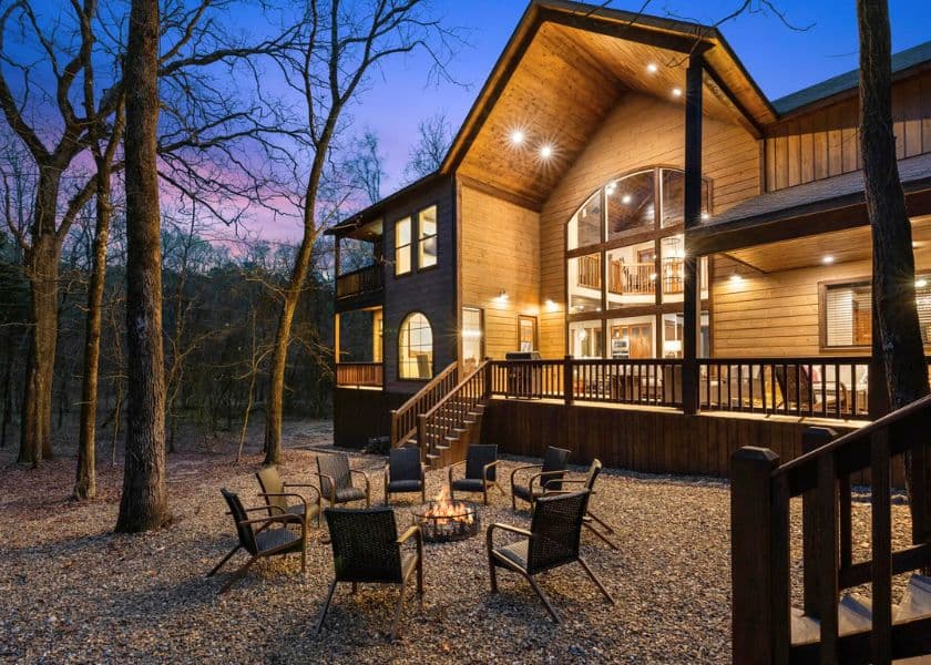 A modern wooden house with large windows, surrounded by trees, featuring a fire pit and seating area in front.