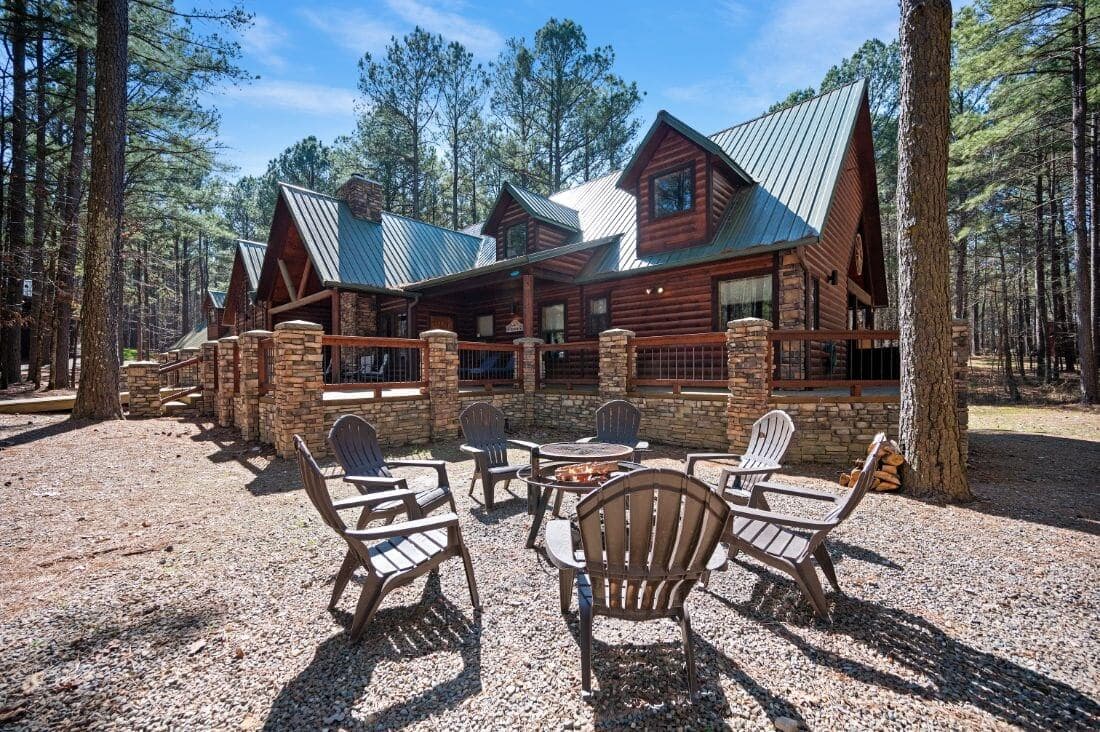 Log cabin surrounded by trees, featuring a fire pit with wooden chairs in the foreground.