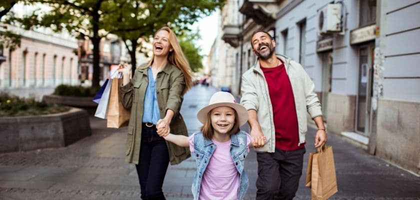 A happy family of three walks hand-in-hand down a city street, carrying shopping bags and smiling.