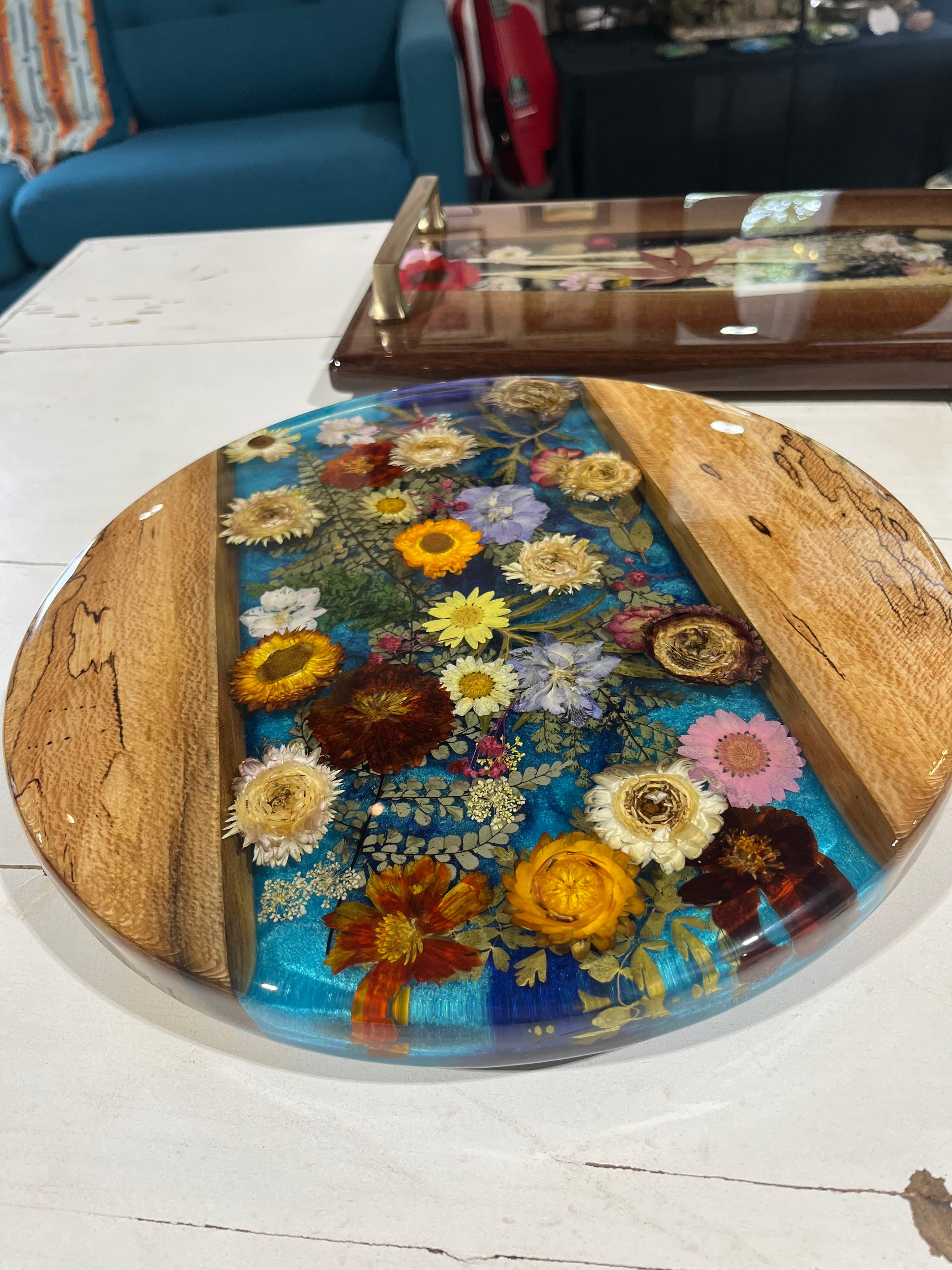 A round resin tabletop showcases colorful dried flowers embedded in a blue background, bordered by wooden edges.
