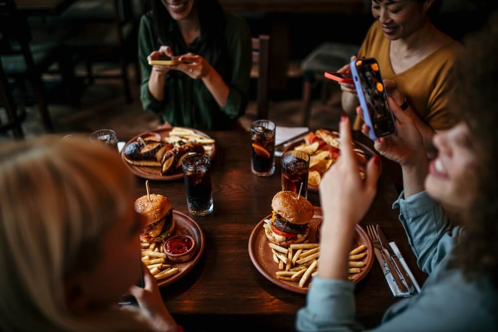 A group of friends sits around a table enjoying burgers and fries, with drinks and phones out for photos. A group of friends sits around a table enjoying burgers and fries, with drinks and phones out for photos.
