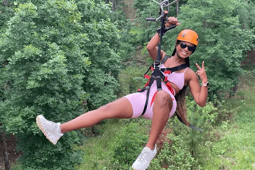 A woman in an orange helmet zips down a zip line, smiling and making a peace sign amidst lush green trees.
