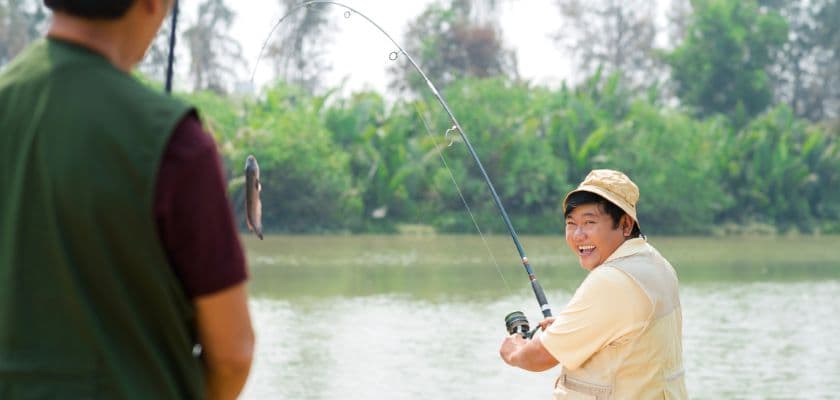 Two people fishing by a pond, one smiling as he reels in a fish.