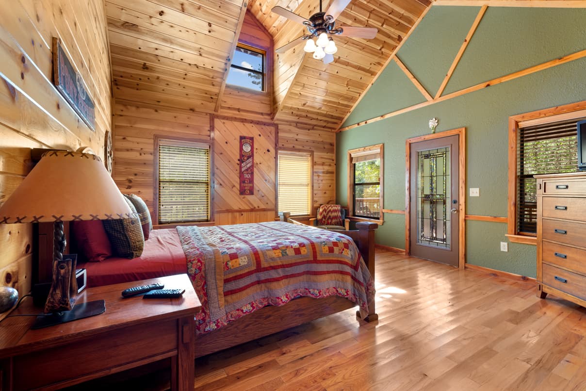 Cozy wooden bedroom with a patterned quilt, ceiling fan, and large windows.