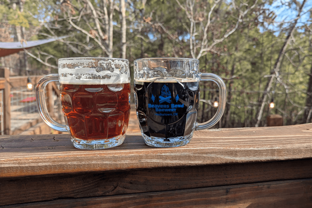 Two glasses of beer, one amber and one dark, resting on a wooden railing amidst a forested background.