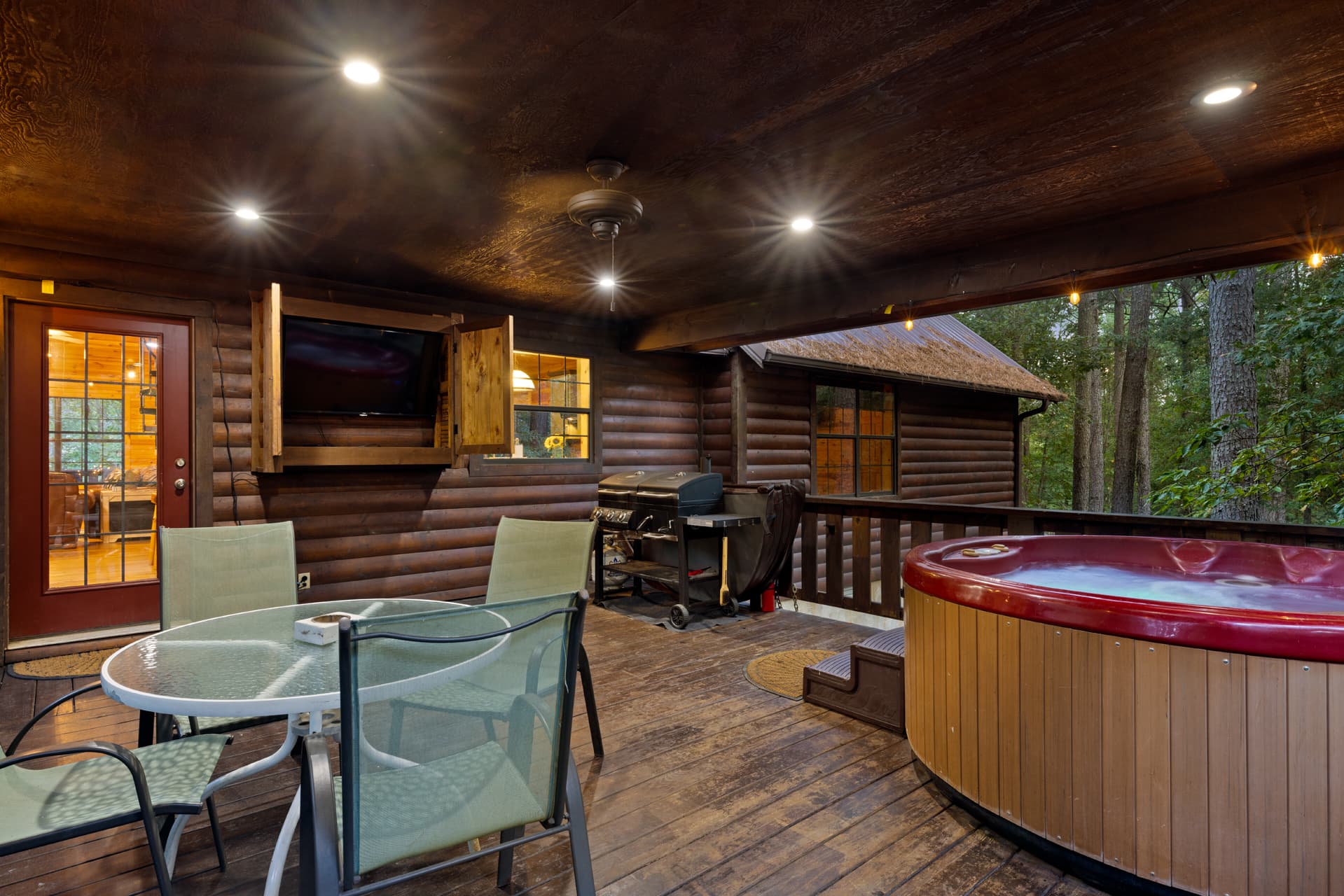 A cozy wooden deck featuring a hot tub, dining table, and outdoor grill, surrounded by trees.